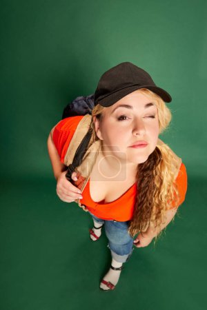 Foto de Portrait of blonde woman in cap with backpack posing over green studio background. Travelling. Vacation time. Concept of american style, culture, emotions, facial expression, lifestyle - Imagen libre de derechos