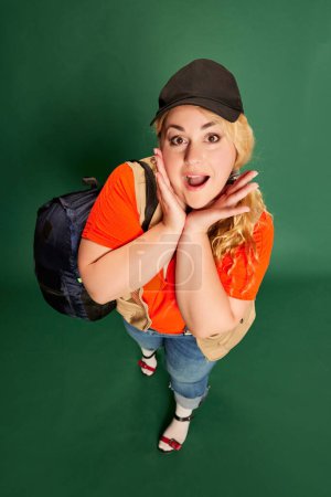 Photo pour Portrait of blonde woman in cap with backpack posing over green studio background. Travelling. Feeling excited . Concept of american style, culture, emotions, facial expression, lifestyle - image libre de droit