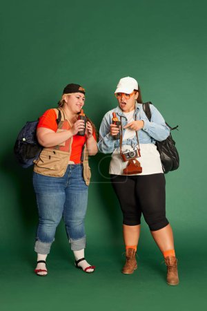Téléchargez les photos : Portrait of two overweight women, friends in casual clothes with backpacks, eating hot dogs over green studio background. Concept of american style, culture, emotions, facial expression, lifestyle - en image libre de droit