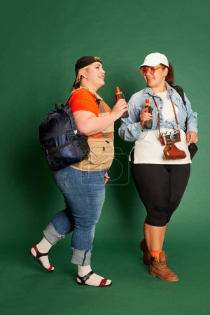 Foto de Portrait of two overweight women, friends in casual clothes with backpacks posing over green studio background. Travel. Concept of american style, culture, emotions, facial expression, lifestyle - Imagen libre de derechos