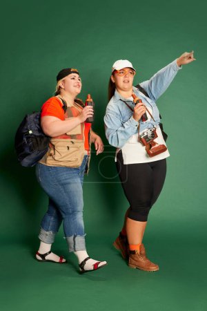 Foto de Portrait of two overweight women, friends in casual clothes with backpacks posing over green studio background. Travelling. Concept of american style, culture, emotions, facial expression, lifestyle - Imagen libre de derechos