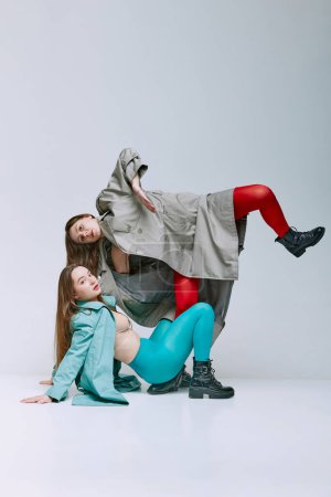 Téléchargez les photos : Portrait of two young women in bright red and blue tights and coat posing over grey studio background. Uniqueness. Concept of modern fashion, queer, art photography, weird people, creativity - en image libre de droit
