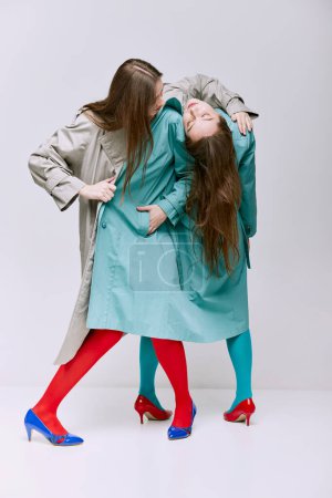 Téléchargez les photos : Portrait of two young girls in bright red, blue tights and coat posing over grey studio background. Concept of modern fashion, queer, art photography, weird people, creativity - en image libre de droit