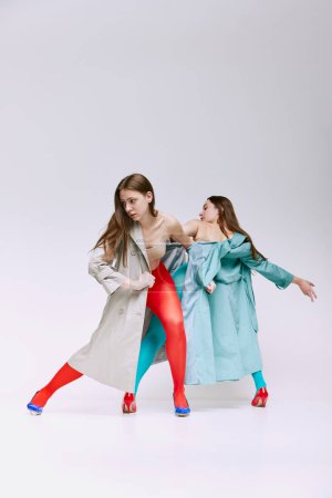 Téléchargez les photos : Portrait of two young girls in bright red and blue tights and coat posing over grey studio background. Inner world. Concept of modern fashion, queer, art photography, weird people, creativity - en image libre de droit