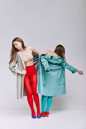 Téléchargez les photos : Portrait of two young girls in bright red and blue tights and coat posing over grey studio background. Modernity. Concept of modern fashion, queer, art photography, weird people, creativity - en image libre de droit