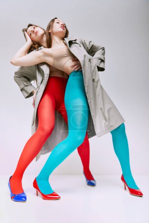 Weirdness and uniqueness. Portrait of two young girls in bright red, blue tights and coat posing on grey studio background. Concept of modern fashion, queer, art photography, weird people, creativity