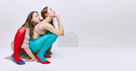 Téléchargez les photos : Portrait of two young girls in bright red and blue tights and coat posing over grey studio background. Banner. Concept of modern fashion, queer, art photography, weird people, creativity - en image libre de droit