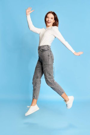 Téléchargez les photos : Feeling happy. Portrait of young beautiful girl in white blouse and jeans posing, jumping over blue studio background. Concept of youth, beauty, fashion, lifestyle, emotions, facial expression. Ad - en image libre de droit