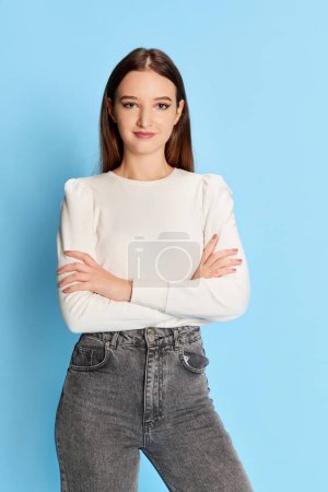 Téléchargez les photos : Portrait of young beautiful girl, student in white blouse and jeans posing over blue studio background. Concept of youth, beauty, fashion, lifestyle, emotions, facial expression. Ad - en image libre de droit
