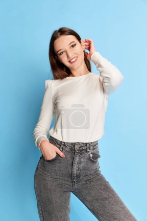 Foto de Cute, smiling. Portrait of young beautiful girl in white blouse and jeans posing over blue studio background. Concept of youth, beauty, fashion, lifestyle, emotions, facial expression. Ad - Imagen libre de derechos