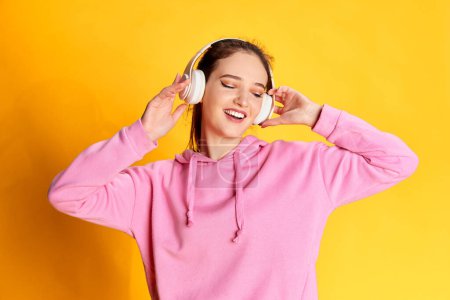 Photo for Portrait of young beautiful girl posing in pink hoodie, listening to music in headphones over yellow studio background. Concept of youth, beauty, fashion, lifestyle, emotions, facial expression. Ad - Royalty Free Image