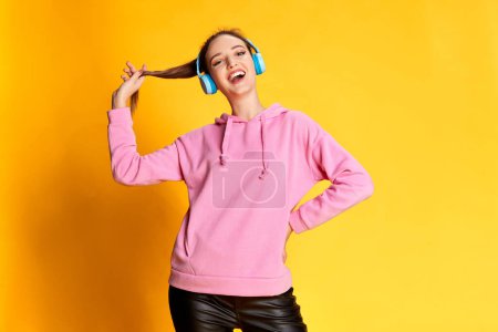 Téléchargez les photos : Portrait of young beautiful girl posing in pink hoodie, listening to music in headphones over yellow studio background. Concept of youth, beauty, fashion, lifestyle, emotions, facial expression. Ad - en image libre de droit