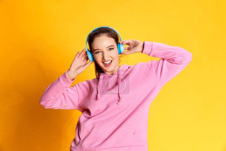 Foto de Portrait of young emotive girl posing in pink hoodie, listening to music in headphones over yellow studio background. Concept of youth, beauty, fashion, lifestyle, emotions, facial expression. Ad - Imagen libre de derechos