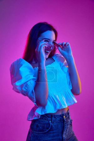 Photo for Portrait of young emotive girl posing in white blouse and trendy sunglasses over purple background in neon light. Concept of youth, beauty, fashion, lifestyle, emotions, facial expression. Ad - Royalty Free Image