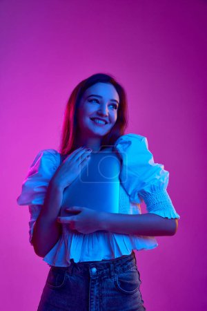 Foto de Portrait of young smiling girl, student in white blouse posing with laptop over purple background in neon light. Concept of youth, beauty, fashion, lifestyle, emotions, facial expression. Ad - Imagen libre de derechos