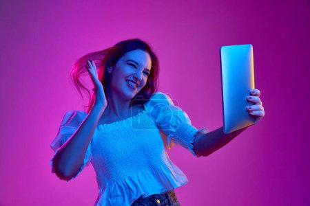 Téléchargez les photos : Portrait of young emotive girl posing in white blouse, haing online video call on tablet on pink background in neon light. Concept of youth, beauty, fashion, lifestyle, emotions, facial expression. Ad - en image libre de droit