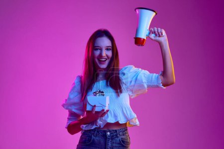 Foto de Portrait of young emotive girl posing with megaphone and present box over pink background in neon light. Holidays. Concept of youth, beauty, fashion, lifestyle, emotions, facial expression. Ad - Imagen libre de derechos