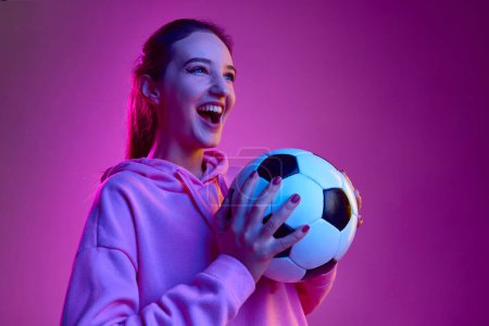 Téléchargez les photos : Emotionally watching match. Portrait of young emotive girl posing with football ball background in neon light. Concept of youth, beauty, fashion, lifestyle, emotions, facial expression. Ad - en image libre de droit
