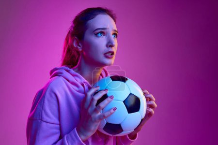 Téléchargez les photos : Tense match moment. Portrait of young emotive girl posing with football ball background in neon light. Concept of youth, beauty, fashion, lifestyle, emotions, facial expression. Ad - en image libre de droit