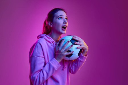 Téléchargez les photos : Tense game, match. Portrait of young emotive girl posing with football ball background in neon light. Concept of youth, beauty, fashion, lifestyle, emotions, facial expression. Ad - en image libre de droit