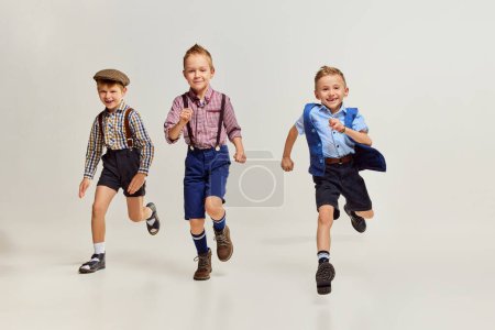 Téléchargez les photos : Active, playful boys, children playing together, running over grey studio background. After school fun. Joy. Concept of game, childhood, friendship, activity, leisure time, retro style and fashion. - en image libre de droit