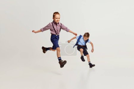 Téléchargez les photos : Joyful boys, children in stylish classical clothes playing together, running, jumping over grey studio background. Concept of game, childhood, friendship, activity, leisure time, retro style, fashion. - en image libre de droit