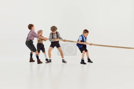 Téléchargez les photos : Motivation to win. Group of little boys, children playing together, pulling the rope over grey studio background. Concept of game, childhood, friendship, activity, leisure time, retro style, fashion. - en image libre de droit