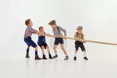 Téléchargez les photos : Having fun. Group of little boys, children playing together, pulling the rope over grey studio background. Concept of game, childhood, friendship, activity, leisure time, retro style, fashion. - en image libre de droit