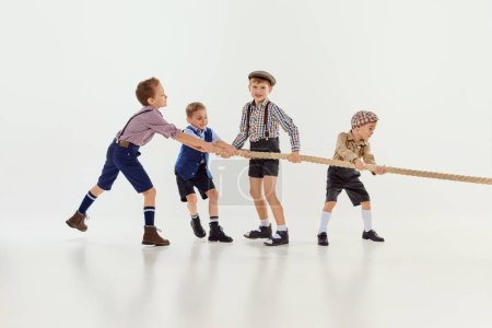 Téléchargez les photos : Sportive game. Group of little boys, children playing together, pulling the rope over grey studio background. Concept of game, childhood, friendship, activity, leisure time, retro style, fashion. - en image libre de droit