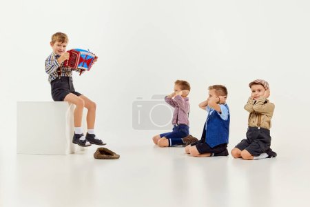 Photo for Boy, child sitting and playing little accordion to his friends covering ears. Children over grey studio background. Concept of game, childhood, friendship, activity, leisure time, retro style, fashion - Royalty Free Image