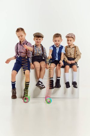 Téléchargez les photos : Group of little boys, children in classical retro clothes sitting and playing with toy over grey studio background. Concept of game, childhood, friendship, activity, leisure time, retro style, fashion - en image libre de droit