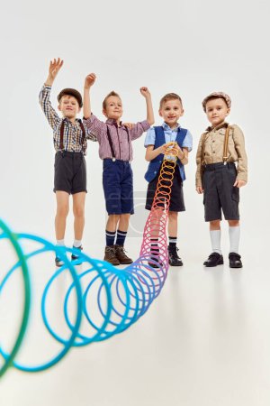 Téléchargez les photos : Boys, children in classical retro clothes playing with slinky toy over grey studio background. Concept of game, childhood, friendship, activity, leisure time, retro style, fashion. - en image libre de droit