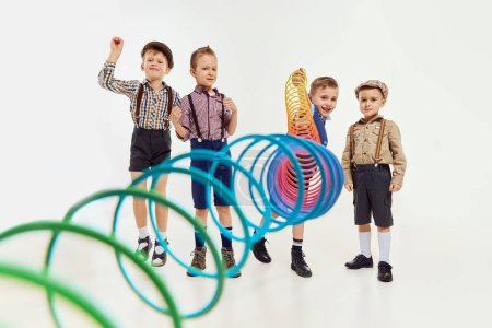 Téléchargez les photos : Group of childen, boys in classical retro clothes playing with slinky toy over grey studio background. Concept of game, childhood, friendship, activity, leisure time, retro style, fashion. - en image libre de droit