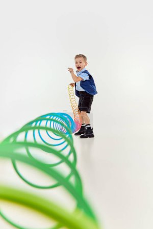 Téléchargez les photos : Happy emotions. Boy, child in classical retro clothes playing with slinky toy over grey studio background. Concept of game, childhood, friendship, activity, leisure time, retro style, fashion. - en image libre de droit