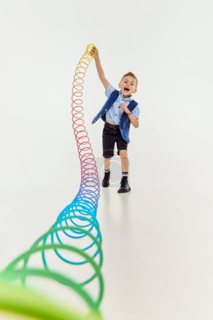 Téléchargez les photos : Joyful game. Boy, child in classical retro clothes playing with slinky toy over grey studio background. Concept of game, childhood, friendship, activity, leisure time, retro style, fashion. - en image libre de droit