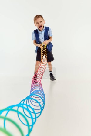 Téléchargez les photos : Boy, child in classical retro clothes playing with slinky toy over grey studio background. Laughing. Concept of game, childhood, friendship, activity, leisure time, retro style, fashion. - en image libre de droit
