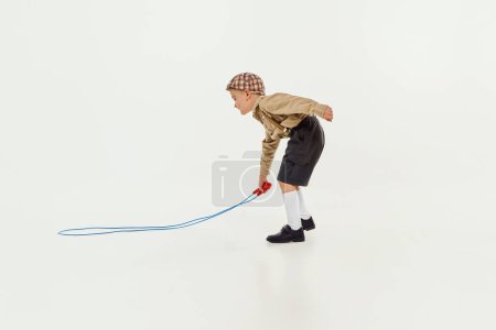Téléchargez les photos : Little boy, child in classical retro clothes playing with jumping rope over grey studio background. Active type of game. Concept of childhood, friendship, activity, leisure time, retro style, fashion. - en image libre de droit
