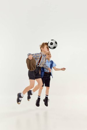 Téléchargez les photos : Hitting ball with head. Boys, children in classical retro clothes playing football over grey studio background. Concept of game, childhood, friendship, activity, leisure time, retro style, fashion. - en image libre de droit