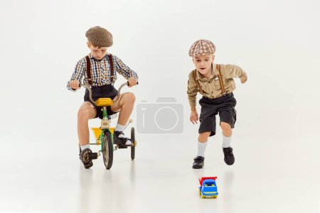 Foto de Active kids. Boys, childred in retro clothes riding vintage bicycle over grey studio background. Concept of game, childhood, friendship, activity, leisure time, retro style, fashion. - Imagen libre de derechos