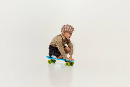 Foto de Little boy, child in classical retro clothes playing, riding skateboard over grey studio background. Concept of game, childhood, friendship, activity, leisure time, retro style, fashion. - Imagen libre de derechos