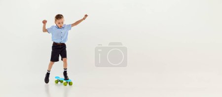 Téléchargez les photos : Playful, active kid, boy riding on skateboard over grey studio background. Banner, flyer. Concept of game, childhood, friendship, activity, leisure time, retro style and fashion. - en image libre de droit