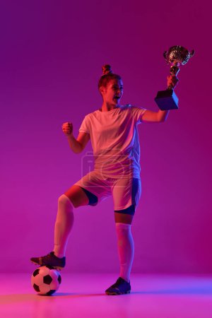 Foto de Young girl, professional female football, soccer player posing with ball and trophy over gradient pink background in neon light. Concept of sport, action, motion, goals, competition, hobby, ad. - Imagen libre de derechos