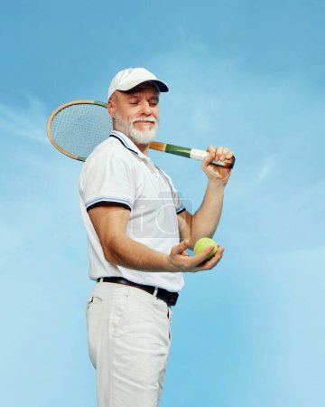 Téléchargez les photos : Country club. Portrait of handsome senior man in stylish white outfit posing with tennis racket over blue background. Concept of leisure activity, hobby, lifestyle, fitness, emotions, retro style - en image libre de droit