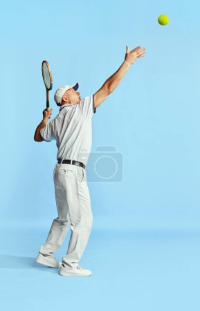 Foto de Serving ball. Portrait of handsome senior man in stylish white outfit playing tennis over blue background. Concept of leisure activity, hobby, lifestyle, fitness, emotions, retro style - Imagen libre de derechos
