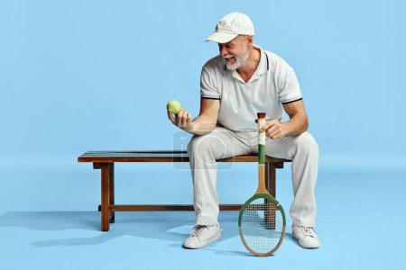 Téléchargez les photos : Portrait of handsome senior man in stylish white outfit sitting on bench with tennis ball and racket over blue background. Concept of leisure activity, hobby, lifestyle, fitness, emotions, retro style - en image libre de droit