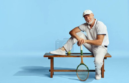 Téléchargez les photos : Portrait of handsome senior man in stylish white outfit sitting on bench over blue background. Tennis country club member. Concept of leisure activity, hobby, lifestyle, fitness, emotions, retro style - en image libre de droit