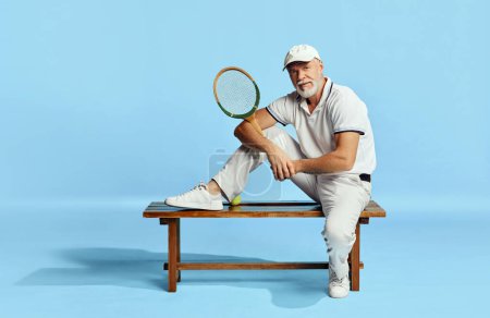 Foto de Popular game. Portrait of handsome senior man in stylish white outfit sitting on bench over blue background. Concept of leisure activity, hobby, lifestyle, fitness, emotions, retro style - Imagen libre de derechos