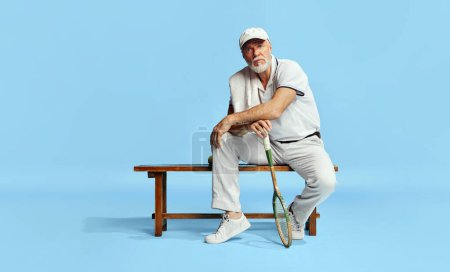 Téléchargez les photos : Portrait of handsome, serious, senior man in stylish white outfit sitting on bench with tennis racket on blue background. Concept of leisure activity, hobby, lifestyle, fitness, emotions, retro style - en image libre de droit