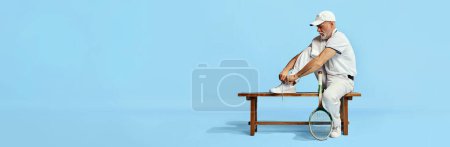 Téléchargez les photos : Portrait of handsome senior man, tennis player in stylish white outfit sitting on bench, tying shoelaces on blue background. Concept of leisure activity, hobby, lifestyle, fitness, retro style. Banner - en image libre de droit