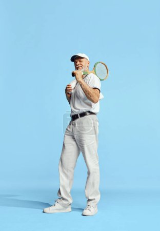 Téléchargez les photos : Noble look. Portrait of handsome senior man in stylish white outfit posing with tennis racket over blue background. Concept of leisure activity, hobby, lifestyle, fitness, emotions, retro style - en image libre de droit